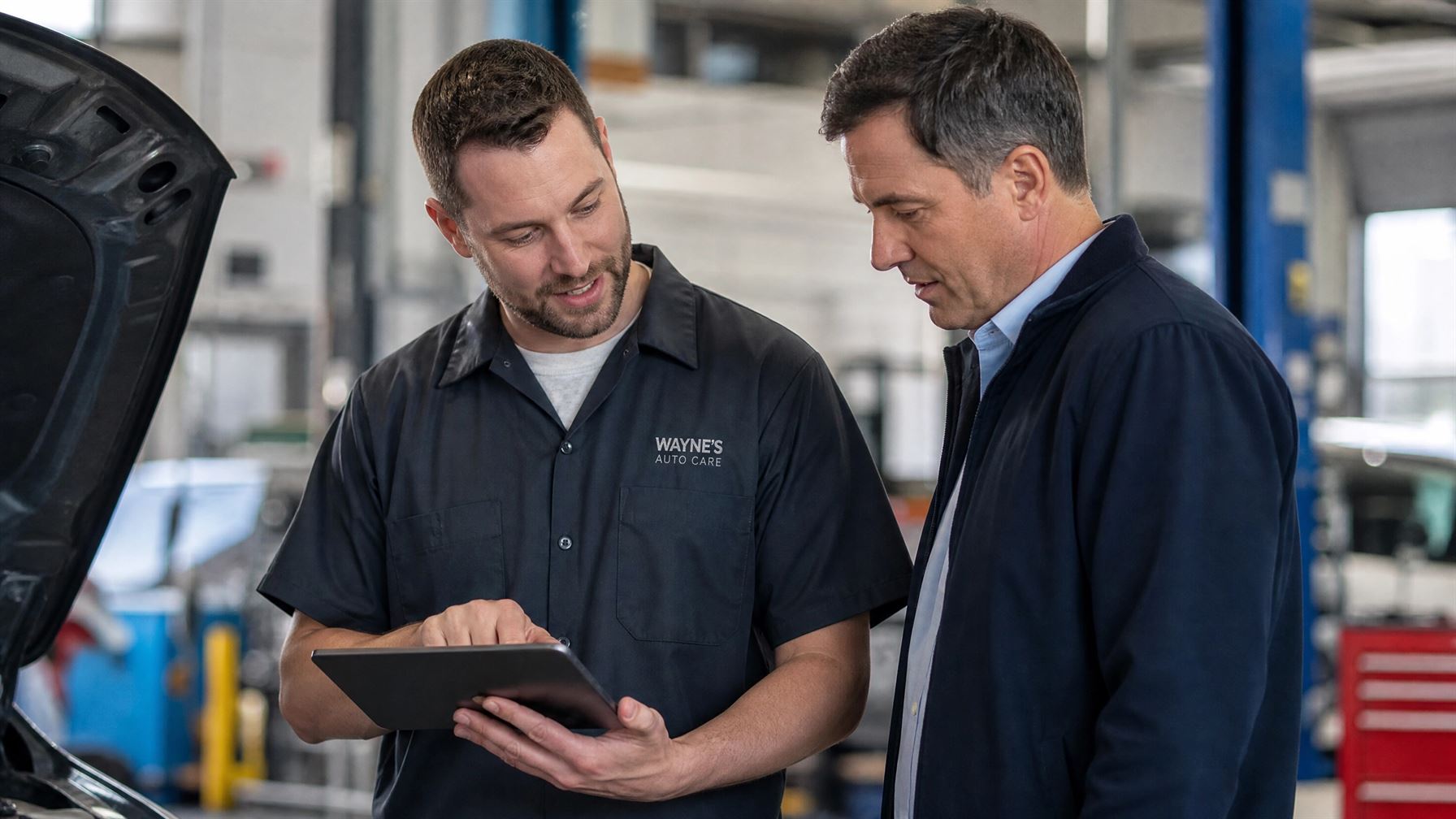 A professional auto mechanic in a clean uniform uses a tablet to explain a vehicle repair estimate to a customer in a modern auto shop.